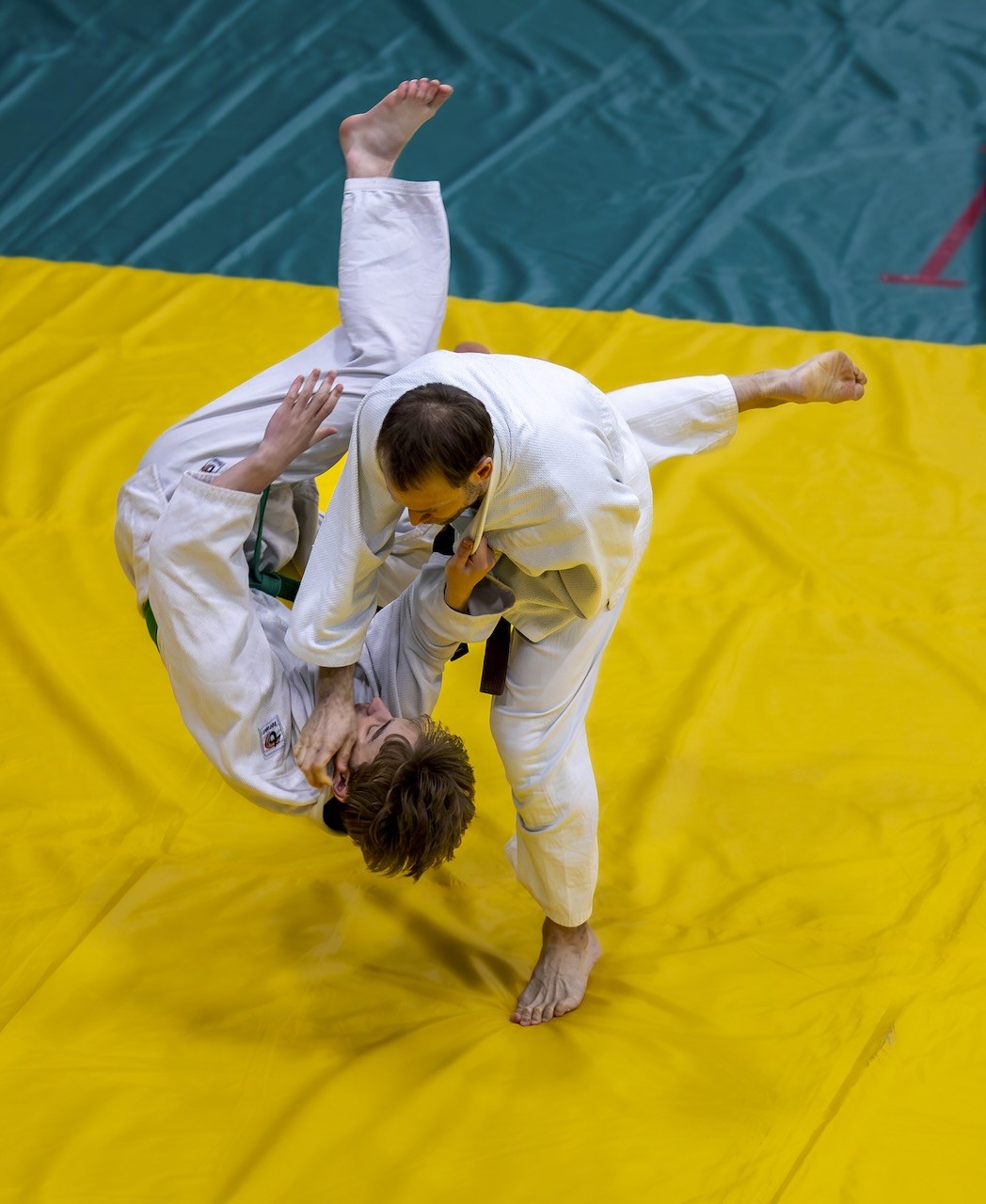 A judoka guiding a partner through a throw on a yellow mat